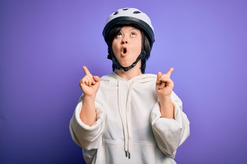 Young down syndrome cyclist woman wearing security bike helmet over purple background amazed and surprised looking up and pointing with fingers and raised arms.