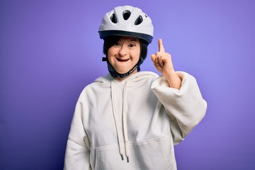 Young down syndrome cyclist woman wearing security bike helmet over purple background pointing finger up with successful idea. Exited and happy. Number one.