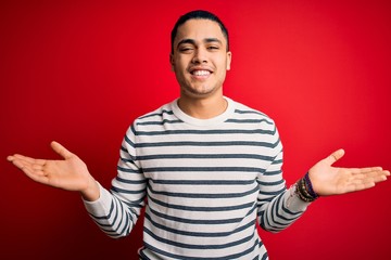 Fototapeta premium Young brazilian man wearing casual striped t-shirt standing over isolated red background smiling showing both hands open palms, presenting and advertising comparison and balance