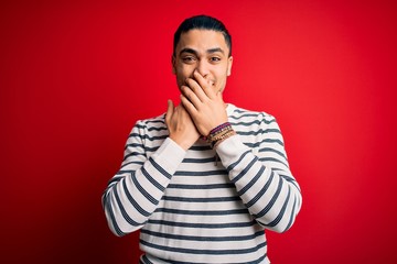 Young brazilian man wearing casual striped t-shirt standing over isolated red background laughing and embarrassed giggle covering mouth with hands, gossip and scandal concept