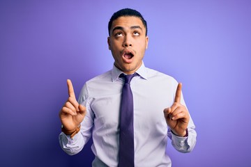 Young brazilian businessman wearing elegant tie standing over isolated purple background amazed and surprised looking up and pointing with fingers and raised arms.