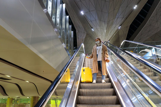 Woman with yellow luggage stands on escalator at almost empty airport terminal due to coronavirus pandemic/Covid-19 outbreak travel restrictions. Flight cancellation. Quarantine measures. 