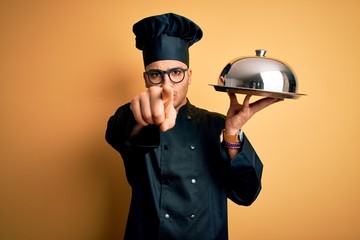 Young brazilian chef man wearing cooker uniform and hat holding tray with dome pointing with finger to the camera and to you, hand sign, positive and confident gesture from the front