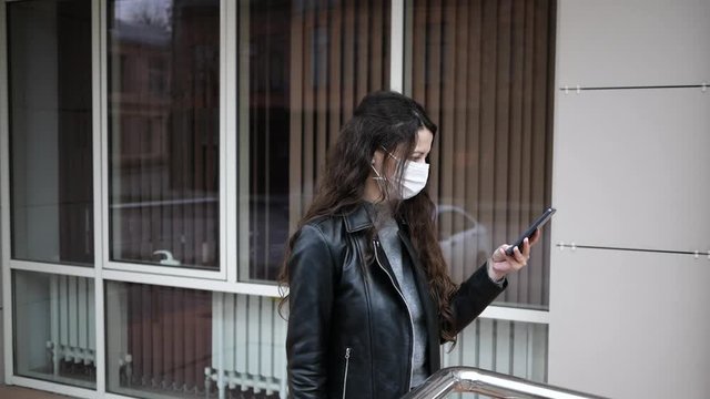 Steadycam shot of caucasian young woman with dark hair out and about in city streets during day, wearing face mask against air pollution and Coronavirus Covid-19, using smartphone