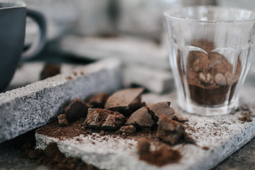 Freshly ground coffee in cup during coffee preparation.Around spilled coffee beans and metal cups with assortment of cookies and nuts on cement brick background.