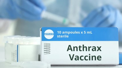 Box with anthrax vaccine on the table against blurred lab assistant. Fictional phaceutical logo