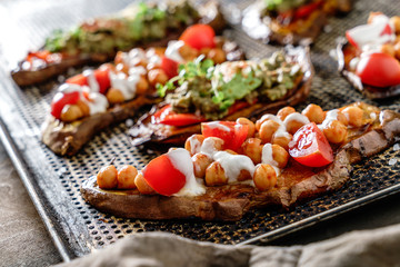 Baked sweet potato toast with roasted chickpeas, tomatoes, goat cheese, sauce guacamole, avocado, seedlings on baking tray over brown background. Healthy vegan food, clean eating, dieting, closeup