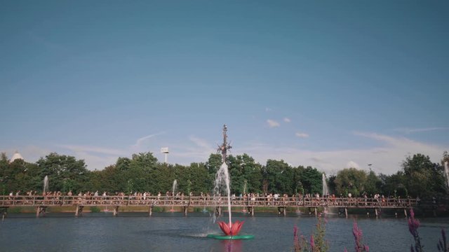 Tomorrowland Belgium connective time lapse of festival guests crossing a bridge accompanied by trees and flower fountains in blue cloudy sky.