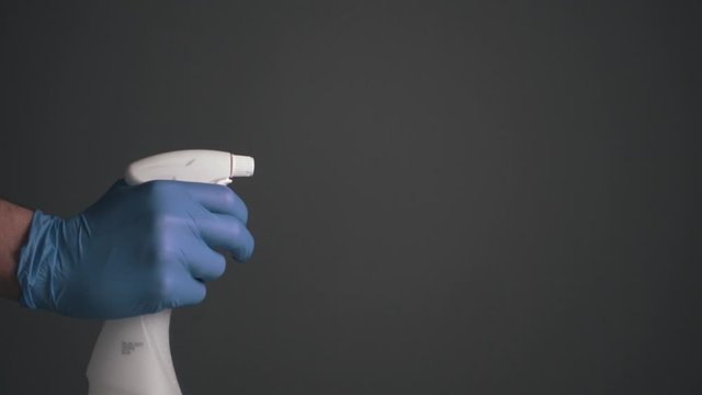 Cinematic, Clean Shot, Dark Background, Male Hand In Blue Plastic Glove Sprays Disinfectant With White Nameless Spray Into Air