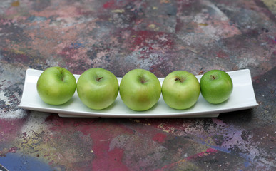 Five organic green apples in a white rectangular bowl on a colorful concrete floor ground