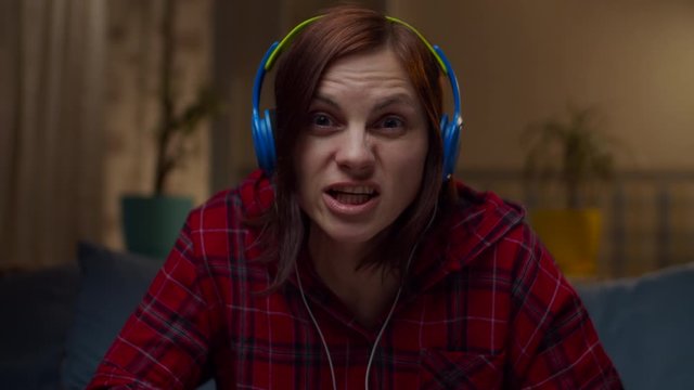 Young 30s woman in red shirt in color headphones talking to camera sitting on sofa at home. Female telling stories looking at camera in slow motion. 