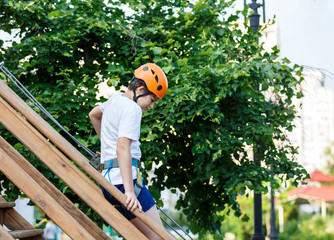 Child in forest adventure park. Kid in orange helmet and white t shirt climbs on high rope trail. Agility skills and climbing outdoor amusement center for children. young boy plays outdoors.