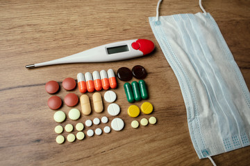 A lot of pills, a body temperature digital thermometer, protective face mask on a wooden table. Medical protective gear and tools. A medications against the COVID-19 virus and other diseases.