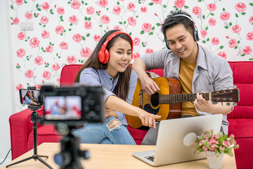 Asian Vlogger of married couple playing and sharing the music to attendee via mobile phone by social media channel,checking comment via laptop, camera taking video and live,vlog and Influencer concept