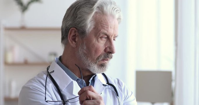 Serious thoughtful old professional doctor looking through window thinking of medical challenge. Concerned senior physician feels worried, pondering on healthcare question, making difficult decision.