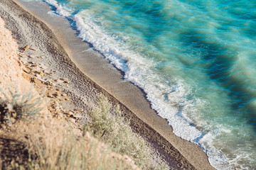 Sunny coastline, beach. View from above on sandy shoreline, blue sea water with white foamy waves. 