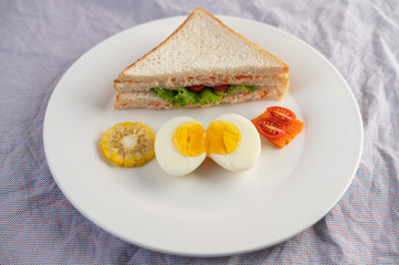 American breakfast on a white background with boiled eggs, corn, tomato and sandwich on a white plate.