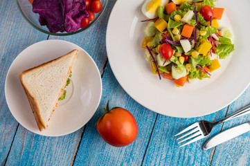 The salad is on a white plate, with a sandwich and tomatoes on the blue wooden floor.