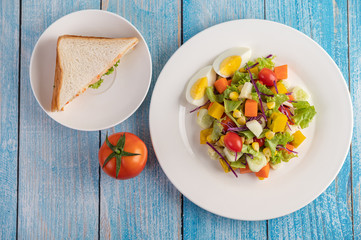 The salad is on a white plate, with a sandwich and tomatoes on the blue wooden floor.