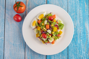 The salad is on a white plate, with a sandwich and tomatoes on the blue wooden floor.