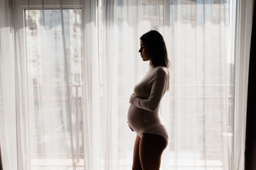 A happy pregnant girl is standing at a large window. strokes the stomach. Baby birth planning concept.