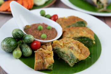 Shrimp paste, along with omelette, Cha-om and eggplant, placed in a white plate on a wooden floor.