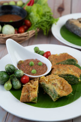 Shrimp paste, along with omelette, Cha-om and eggplant, placed in a white plate on a wooden floor.
