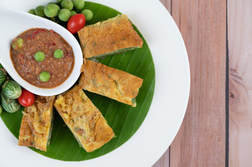 Shrimp paste, along with omelette, Cha-om and eggplant, placed in a white plate on a wooden floor.