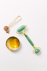 Jade roller, scoop of herbal facial powder in spoon, and oil serum in brass dish on white surface / Holistic wellness and self care concept