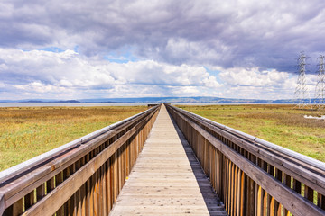 Obraz premium Wooden boardwalk going over the marshes of south San Francisco bay area; electricity towers and lines visible against the cloudy sky; Palo Alto Baylands Park, California