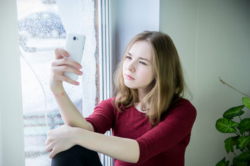 A teenage girl with long hair sits on a windowsill and looks at the screen of a mobile phone, photographs herself, communicates via video communication. It is raining outside the window.
