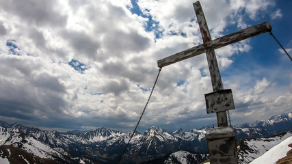 Wooden cross at the top of Himmeleck in Austrian Alps. There is a massive mountain range in the back, partially covered with snow. Early spring vibes. barren mountain slopes. Overcast. Achievement