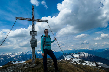 A woman with big hiking backpack standing on the top of Himmeleck in Austrian Alps. There is a massive mountain range in the back, partially covered with snow. Early spring vibes. Cross on the top