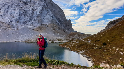 A woman in hiking outfit standing by the Wolayer Lake in Austrian Alps. There is massive, rocky mountain on the other side of the lake. New day beginning. Soft reflections in the lake. Happiness