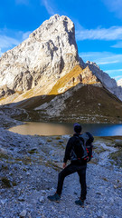 Fototapeta premium A man in hiking outfit enjoying the view on Wolayer Lake in Austrian Alps. There is massive, rocky mountain on the other side of the lake. New day beginning. Soft reflections in the lake. Happiness