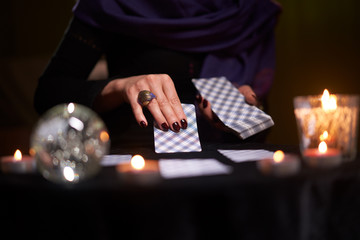 Close-up of female fortuneteller's hands with cards at table with candles, magic ball in dark room