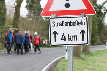 Eine Gruppe Menschen steht auf einer Stra&szlig;e zum Bo&szlig;eln, Verkehrsschild mit Aufschrift Stra&szlig;enbo&szlig;eln 4km