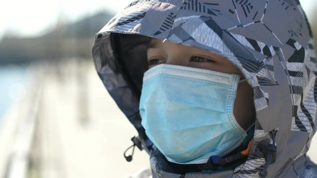 Small boy in a sanitary mask and hood standing in a park on sunny day in spring