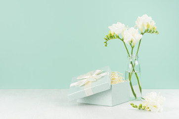 Wedding festive background with opened gift box with paper filler, fragrance soft white flowers freesia  in  vase on green mint menthe wall on white board.