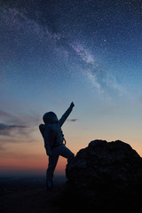 Silhouette of space traveler standing on rocky mountain and pointing at stars, Milky way. Cosmonaut...