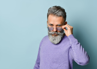 Gray-haired aged man in purple sweater. Fixing his sunglasses and looking at you while posing against blue background. Close up