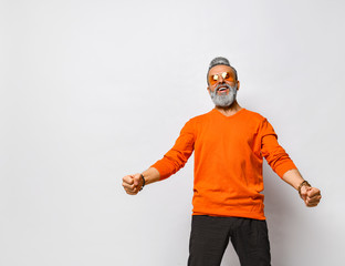 Elderly man in orange sweater, sunglasses, black pants, bracelets. Raised fists , screaming, posing isolated on white. Close up