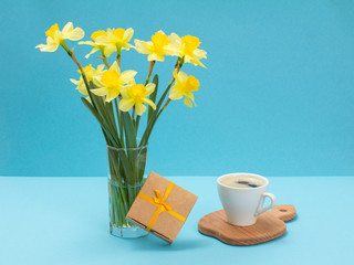 Bouquet of yellow daffodils in vase with gift box on a blue background.