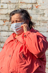 Senior woman stands in a medical mask on her face, which protects against coronavirus and other viruses and diseases on a background of old brick wall. Coronavirus pandemic