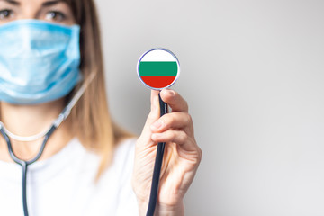 female doctor in a medical mask holds a stethoscope on a light background. Added flag of Bulgaria....