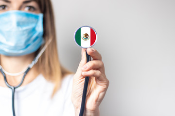 female doctor in a medical mask holds a stethoscope on a light background. Added flag of Mexico....