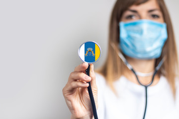 female doctor in a medical mask holds a stethoscope on a light background. Added flag of Canary Islands. Concept medicine, level of medicine, virus, epidemic