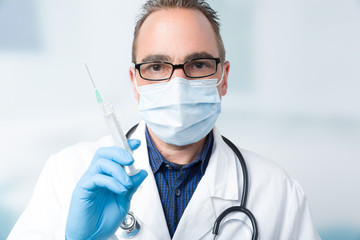 male doctor with medical face mask and medical gloves holding a syringe in front of a clinic room