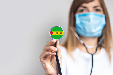 female doctor in a medical mask holds a stethoscope on a light background. Added flag of Tome and...