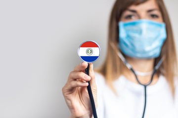 female doctor in a medical mask holds a stethoscope on a light background. Added flag of Paraguay....